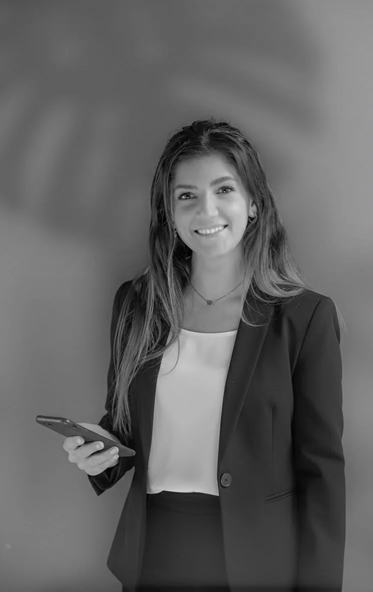 Beatrice Pirani in piedi davanti a una parete chiara, giacca scura e telefono in mano.